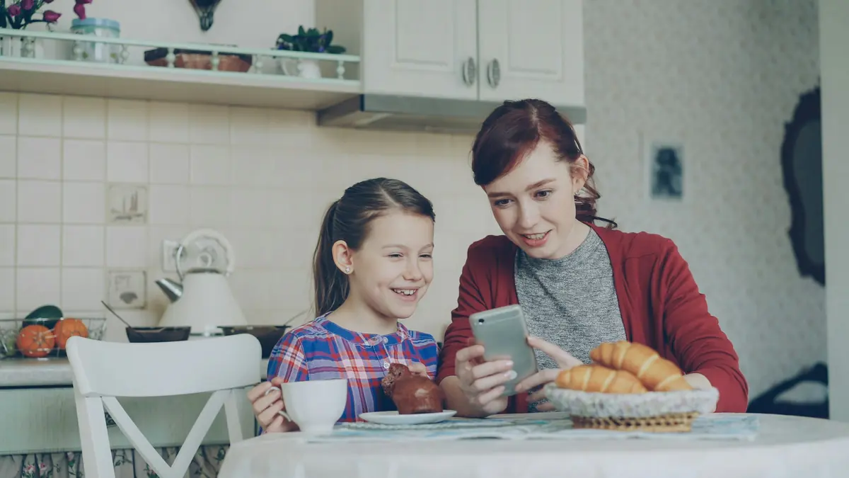 En mor og en datter ser på en mobiltelefon sammen ved frokostbordet